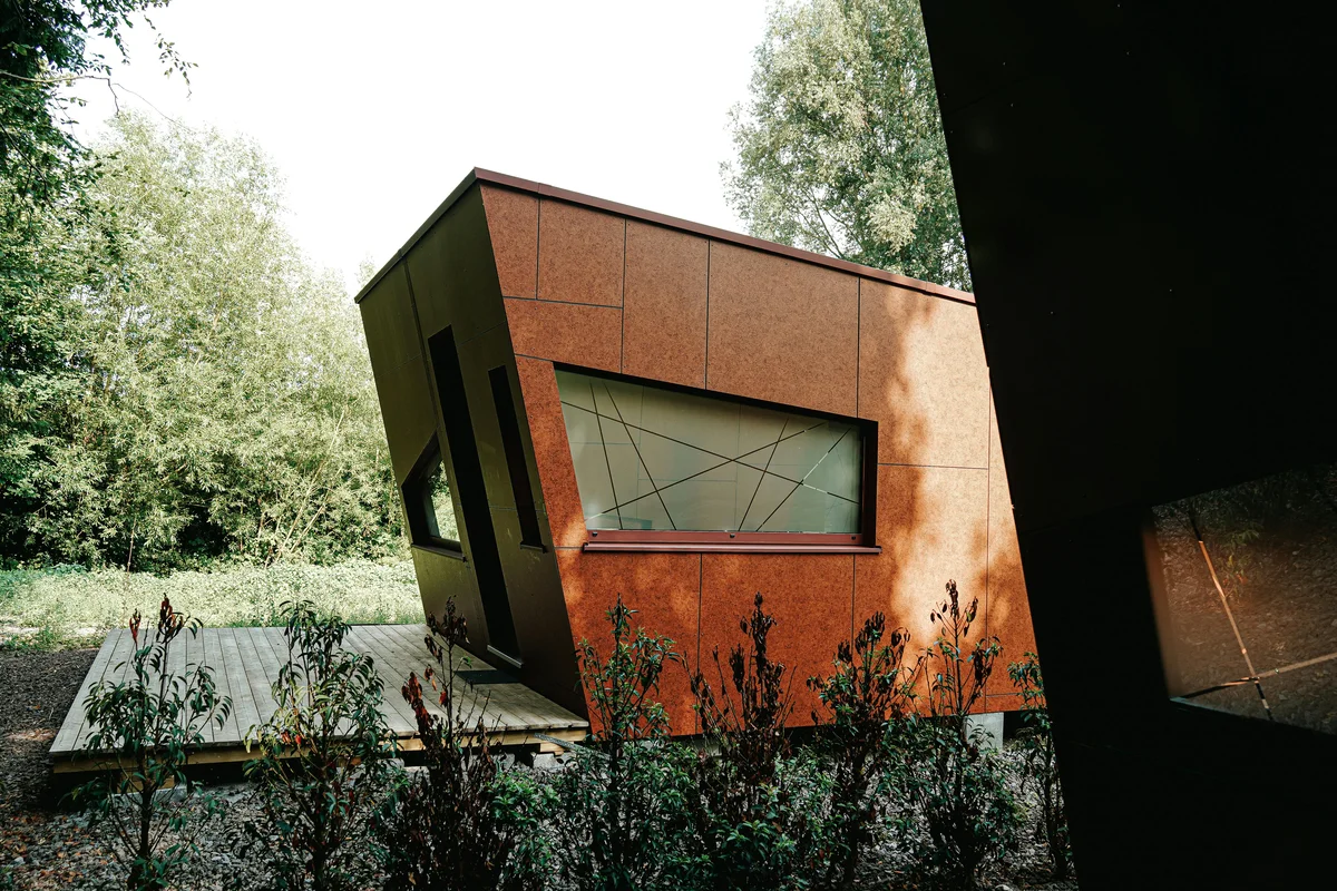 Cabană mică cu fațadă din tablă Corten patinată — exterior brut-industrial integrat în peisaj natural verde